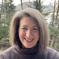 Profile picture of Sheri Von Wolfe.  She has short brown hair, green eyes, and is smiling.  She is standing in front of trees and a lake.