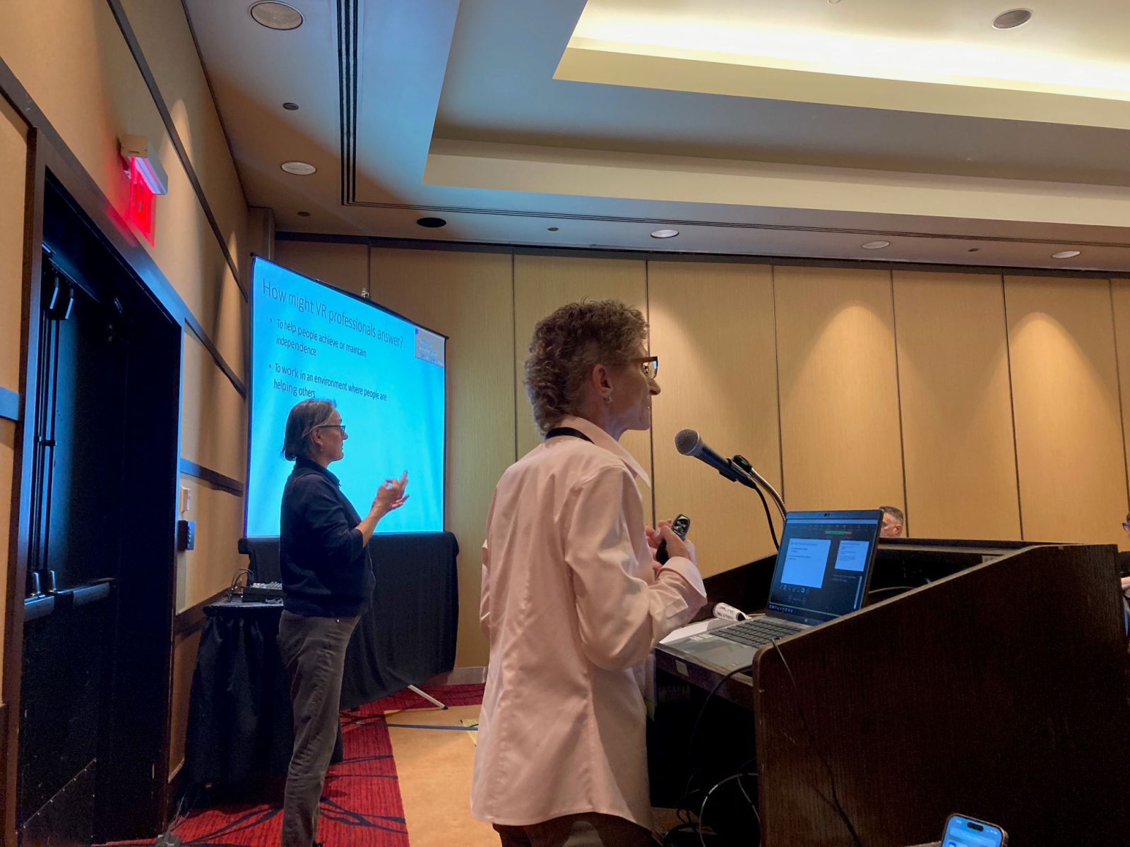 Kelly standing at podium in meeting room with projected slide and ASL interpreter in background.