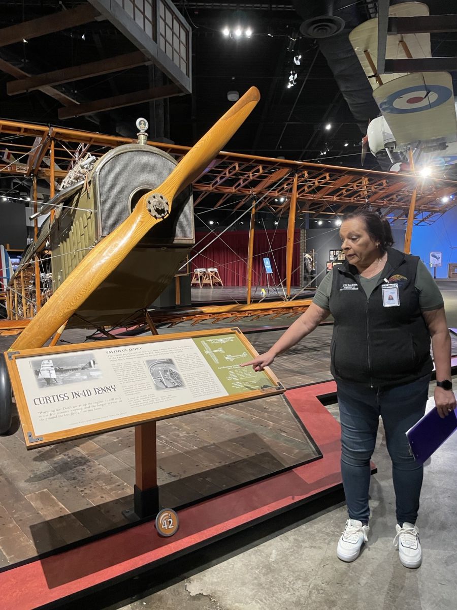 Woman stands in front of an antique airplane and points to a exhibit sign titled Curtiss JN-4D Jenny