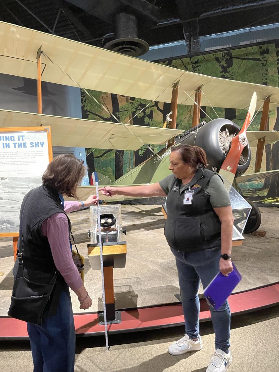 In front of an antique plane, 2 women measure a post-mounted clear box containing an interactive viewing device