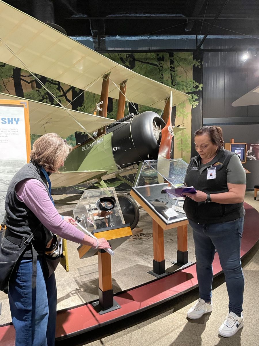 In front of an antique plane, a woman measures a clear plastic display case while another woman writes on a clipboard