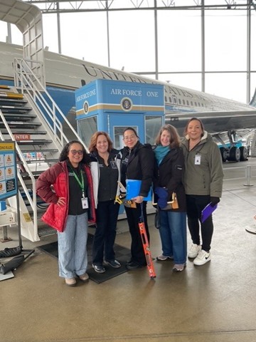 A group of five women of different ethnicities smile in front of an Air Force One plane.