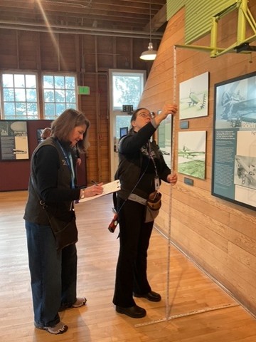 Two women with light skin tone measure the height of a wall-mounted metal airplane part and take notes on a clipboard in a gallery with walls and floor made of wood.